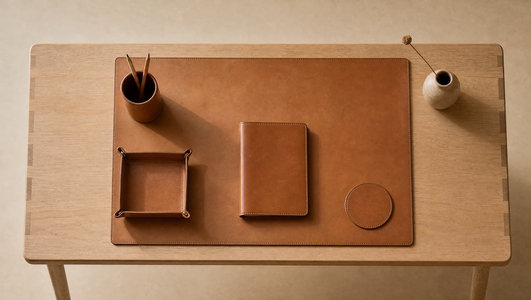 A writing desk seen from above, fully dressed in leather: mat, pen cup, valet tray, notebook cover and a coaster on a pale oak top